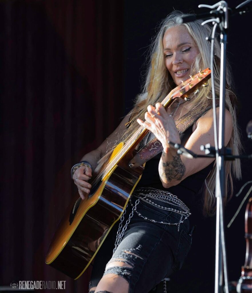 A woman with long blonde hair plays an acoustic guitar on stage, wearing ripped black jeans and a chain belt.